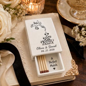 May include: A white rectangular ceramic matchbox with a sliding drawer containing wooden matches. The top features a black line drawing of a cherub pouring liquid into a fountain, with the text Olivia & Daniel 06.21.2026. The interior reads A Perfect Match.
