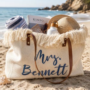 Könnte beinhalten: Eine cremefarbene, gewebte Tragetasche mit braunen Ledergriffen und Fransenbesatz, die den gestickten dunkelblauen Text "Mrs. Bennett" aufweist. Die Tasche enthält ein blau-weiß gestreiftes Handtuch, ein Kinfolk-Magazin, eine Sonnenbrille, eine Flasche und einen Strohhut.