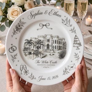 May include: A white ceramic decorative plate featuring a black ink illustration of a castle, palm trees, and a pond. The text reads Sophia & Ethan, The White Castle, Montecito, California, and June 15, 2026. Accents include wedding rings, doves, a cake, and champagne flutes.
