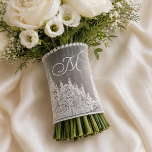 Op de afbeelding: Een boeket witte rozen en gipskruid met groene stelen, omwikkeld met een doorschijnend wit lint. Het lint is voorzien van een wit borduursel van een kathedraal en een cursief geborduurde letter M als monogram in witte draad.