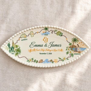 May include: An oval ceramic tray with a beaded edge featuring a dotted travel path connecting illustrations of NYC, Australia, Miami, and Honolulu. The center displays the text "Emma & James", "Can't Help Falling in Love", and "November 7, 2026" in gold and black script.