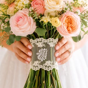 Peut inclure: Un bouquet floral composé de roses rose pâle, pêche et crème avec du feuillage vert. Les tiges sont enveloppées dans un large ruban de dentelle blanche avec un bord festonné complexe et une lettre M blanche brodée au centre.