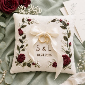 May include: A square off-white ring pillow featuring embroidered dark red roses and green foliage. A cream satin ribbon is tied in a bow at the center, holding two gold rings. The text S & L and the date 10.24.2026 are embroidered in brown thread.