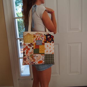 May include: A patchwork tote bag featuring a variety of autumn-themed fabric squares in shades of orange, yellow, brown, and green. The bag includes a central blue rectangular label, cream-colored fabric handles, and a decorative white lace trim.