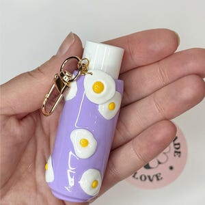 May include: A light purple cylindrical lip balm holder featuring raised, three-dimensional fried egg motifs with white whites and yellow yolks. The holder includes a gold-toned metal lobster clasp keychain attached to the top white cap section.