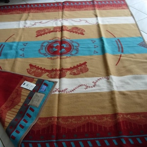 May include: A rectangular blanket featuring horizontal stripes in tan, white, and red with a central turquoise band. The design includes red circular motifs with bear paw prints, animal silhouettes, and geometric patterns. A corner is folded back.