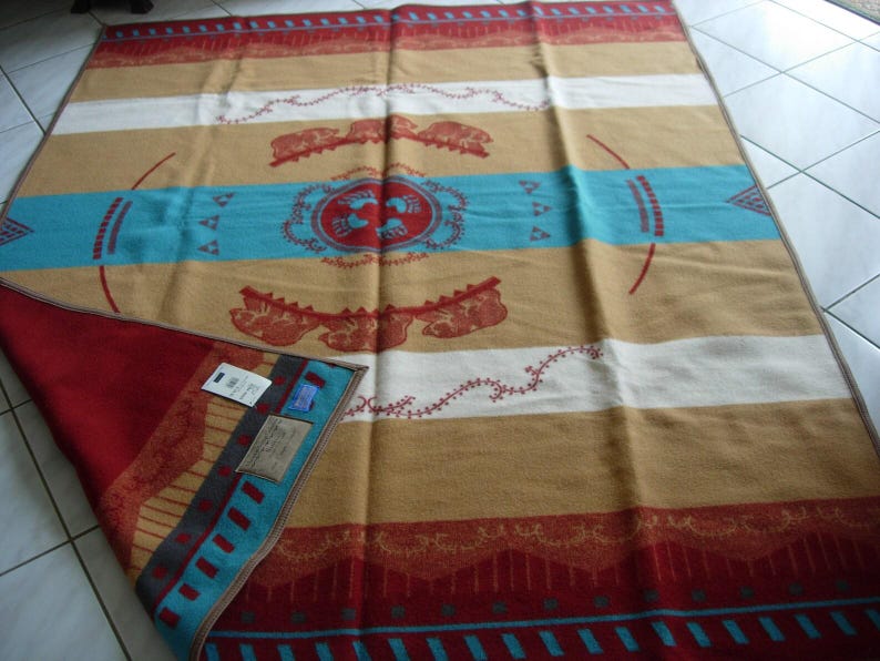 May include: A rectangular blanket featuring horizontal stripes in tan, white, and red with a central turquoise band. The design includes red circular motifs with bear paw prints, animal silhouettes, and geometric patterns. A corner is folded back.
