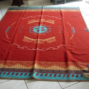 May include: A large rectangular blanket in a deep red hue featuring a central light blue circular emblem with bear paw prints. The design includes tan and turquoise geometric borders, stylized animal motifs, and delicate white line patterns.