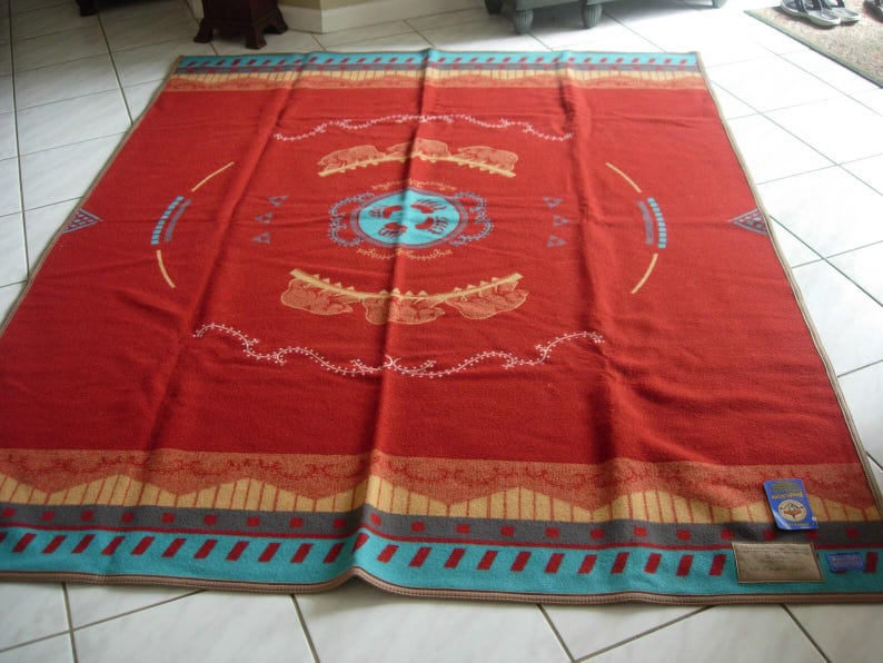 May include: A large rectangular blanket in a deep red hue featuring a central light blue circular emblem with bear paw prints. The design includes tan and turquoise geometric borders, stylized animal motifs, and delicate white line patterns.