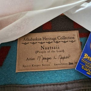 May include: A Pendleton Beaver State wool blanket from the Athabaskan Heritage Collection titled Naatsaii. The blanket features a teal and brown geometric pattern with a tan fabric label and a blue product tag detailing the wool and cotton blend.