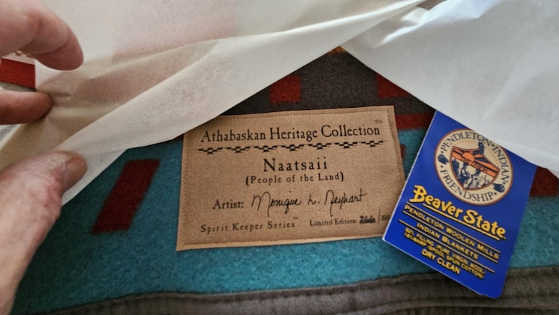 May include: A Pendleton Beaver State wool blanket from the Athabaskan Heritage Collection titled Naatsaii. The blanket features a teal and brown geometric pattern with a tan fabric label and a blue product tag detailing the wool and cotton blend.