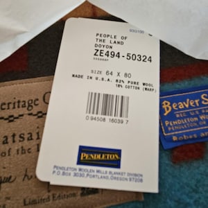 May include: A white product tag with black text reading People of the Land Doyon, ZE494-50324, size 64 x 80, made in U.S.A., 82% pure wool, 18% cotton, and a barcode. Beside it are a tan Athabaskan Heritage label and a blue Beaver State Pendleton label.