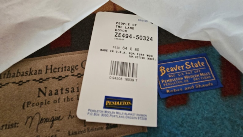 May include: A white product tag with black text reading People of the Land Doyon, ZE494-50324, size 64 x 80, made in U.S.A., 82% pure wool, 18% cotton, and a barcode. Beside it are a tan Athabaskan Heritage label and a blue Beaver State Pendleton label.