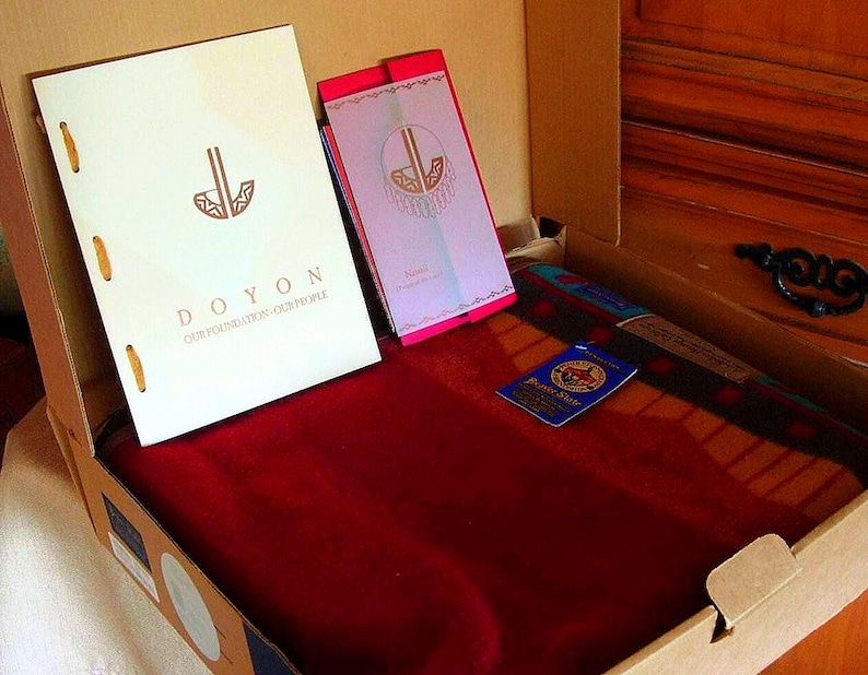May include: A cardboard box containing a white booklet with the text DOYON OUR FOUNDATION OUR PEOPLE, a smaller pink and white booklet, a blue tag with a circular logo, and a folded deep red textile with a patterned border in a box.
