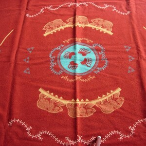 May include: A red woven blanket featuring a central light blue circle with four bear paw prints. The design includes two horizontal rows of three bears, decorative white scrollwork borders, and geometric patterns in light blue and yellow.