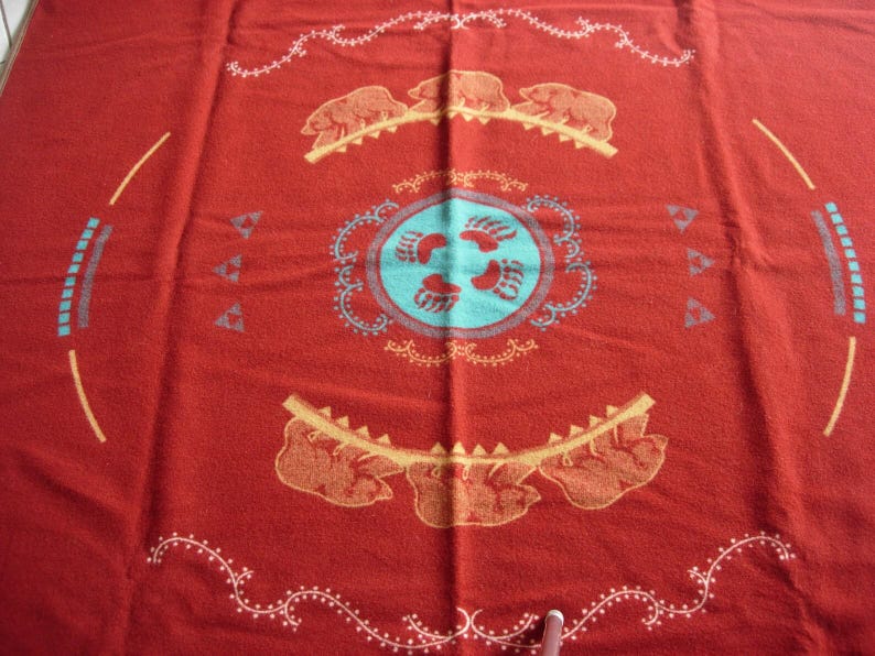 May include: A red woven blanket featuring a central light blue circle with four bear paw prints. The design includes two horizontal rows of three bears, decorative white scrollwork borders, and geometric patterns in light blue and yellow.