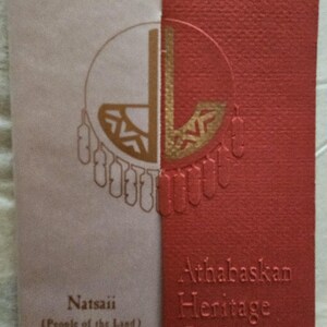 May include: A rectangular card with a textured red and light beige surface. The red section features the embossed text Athabaskan Heritage Collection. The beige section displays the text Natsaii (People of the Land) and a gold circular graphic.