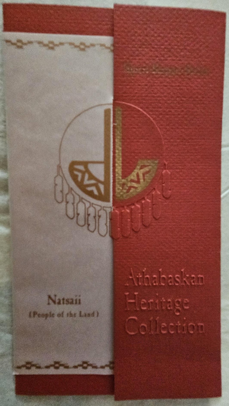 May include: A rectangular card with a textured red and light beige surface. The red section features the embossed text Athabaskan Heritage Collection. The beige section displays the text Natsaii (People of the Land) and a gold circular graphic.