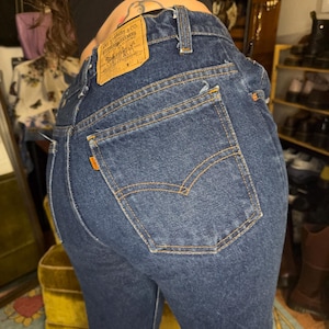 May include: Dark blue denim jeans featuring a high-waisted fit, a tan leather patch on the back waistband with the text Levi Strauss & Co., and a small orange tab on the back pocket. The jeans have visible orange stitching and belt loops.