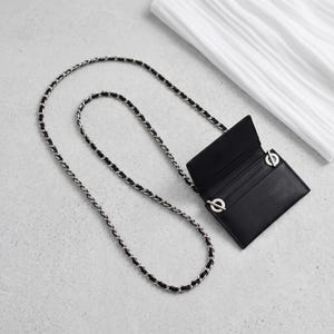 May include: A black rectangular card holder with an open flap and multiple interior slots. The accessory features a long, thin shoulder strap made of a silver-toned metal chain interwoven with a black cord, attached by two circular silver rings.