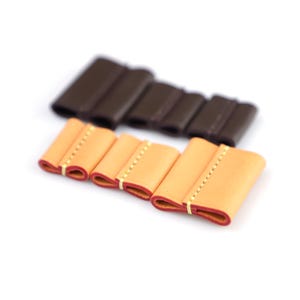May include: A set of seven leather cord organizers arranged in two rows. The front row features three light tan leather pieces with red edges and yellow stitching. The back row contains four dark brown leather pieces with matching stitching.