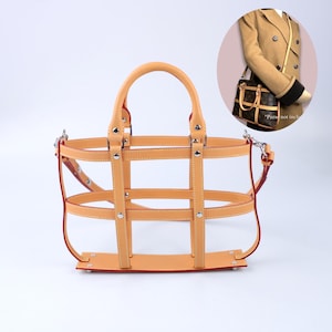 Puede incluir: Un organizador de bolso de cuero color canela claro estilo jaula con un patrón de rejilla de correas verticales y horizontales, remaches de metal plateado y una base rectangular plana. Incluye dos asas superiores y una correa de hombro.