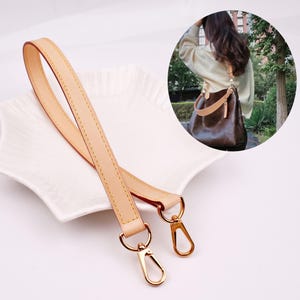 May include: A light tan leather replacement bag strap featuring gold-tone metal lobster claw clasps at both ends. The strap is long and slender with visible stitching along the edges. An inset shows the strap attached to a dark brown handbag.