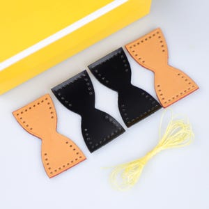 Puede incluir: Cuatro piezas de cuero en forma de reloj de arena con agujeros preperforados en los bordes, incluyendo dos piezas color canela y dos negras, acompañadas de un manojo de hilo amarillo fino. Las piezas están alineadas junto a un objeto amarillo.