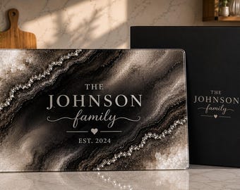May include: A rectangular glass cutting board featuring a black and white marbled geode pattern with silver glitter accents. The text reads THE JOHNSON family EST. 2024 with a small heart. It is displayed next to a matching black gift box.