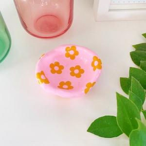 May include: A small pink ceramic dish with a yellow daisy pattern. The dish is ideal for holding jewellery or other small items.
