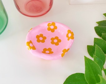 May include: A small pink ceramic dish with a yellow daisy pattern. The dish is perfect for holding jewelry or other small items.