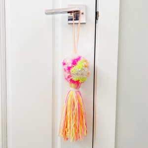 Peut inclure: Un porte-clé en pompon coloré avec un long gland en fil rose, jaune et blanc. Le porte-clé est accroché à une porte blanche avec une poignée de porte argentée.