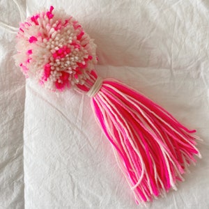 May include: A vibrant yarn pom-pom and tassel in pink and white. The pom-pom is a fluffy ball of white yarn with pink flecks. The tassel features pink and white yarn strands, bound with a natural string, creating a decorative item.