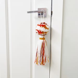 Peut inclure: Un pompon décoratif suspendu à une poignée de porte. Le pompon est composé de fil blanc, orange et marron. La gland en dessous est blanc, orange et or. La porte est blanche.