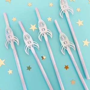 May include: Set of six iridescent silver straws with white rocket ship cutouts. The straws are on a light blue background with gold glitter stars.