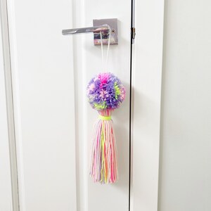 May include: A vibrant yarn tassel door knob hanger. The top is a pom-pom in shades of purple, pink, and green. The tassel is made of long strands of yarn in pink, white, and yellow. A yellow yarn wraps the tassel base. The hanger is attached to a silver door handle.