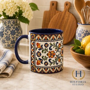 May include: A ceramic mug featuring a dark blue handle and interior, decorated with a vibrant, symmetrical floral pattern in shades of orange, yellow, green, and blue against a white background, framed by ornate, colorful geometric borders.