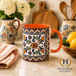 May include: A ceramic mug featuring a vibrant, multi-colored floral pattern with orange, blue, and green hues on a white background. The mug has a bright orange interior and a matching orange handle, with a decorative border design at the top and bottom.