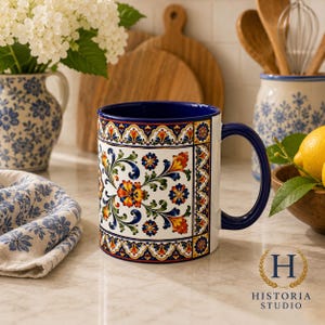 May include: A ceramic mug featuring a vibrant, multi-colored floral pattern with orange, red, green, and blue hues on a white background. The mug has a deep blue interior and a matching blue handle, with a decorative border design.