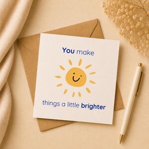 May include: A square white greeting card featuring a yellow sun illustration with a simple black smiley face. The text reads "You make things a little brighter" in blue lettering. The card is placed on a brown envelope with a pen nearby.