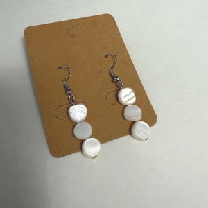 May include: A pair of drop earrings featuring three white, iridescent, circular shell beads suspended from silver-toned metal fishhook wires. The earrings are attached to a rectangular brown cardboard display card with a circular hole at the top.