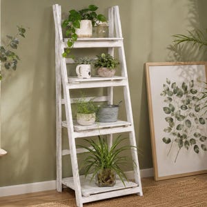 May include: A white wooden ladder shelf displaying various potted plants and decorative items. The shelf is against a light green wall, with a framed botanical print and a woven basket nearby. The plants include succulents and ferns.