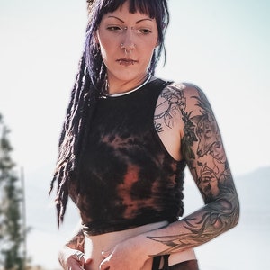 May include: A woman with purple hair and tattoos is wearing a black tie-dye crop top and matching shorts. The outfit has a tie detail at the waist. She is standing in a field of tall grass.