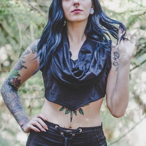 May include: A woman with blue hair and tattoos wears a black crop top with a cowl neck and a long black skirt with side slits. The skirt has a lace-up detail at the waist.