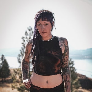May include: A person with purple hair and tattoos is wearing a black tie-dye crop top and matching black tie-dye shorts. They are standing in a field with a body of water in the background.