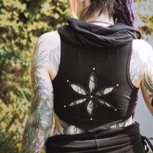 May include: A black sleeveless top with a cut-out design in the shape of a flower. The top has a hood and is worn by a person with tattoos on their arms.