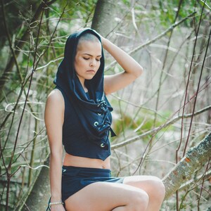 May include: A woman wearing a black hooded top with a lace-up front and a short black skirt. She is sitting on a tree branch with her legs crossed and wearing gray knee-high boots.
