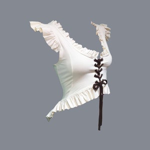 May include: A white crop top with a lace-up front and ruffled trim. The top has a brown ribbon tied at the front.