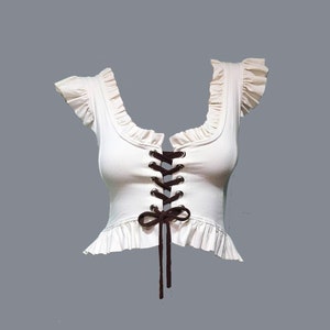 May include: A white crop top with a lace-up front and a ruffled neckline. The top has brown laces that tie into a bow at the centre.