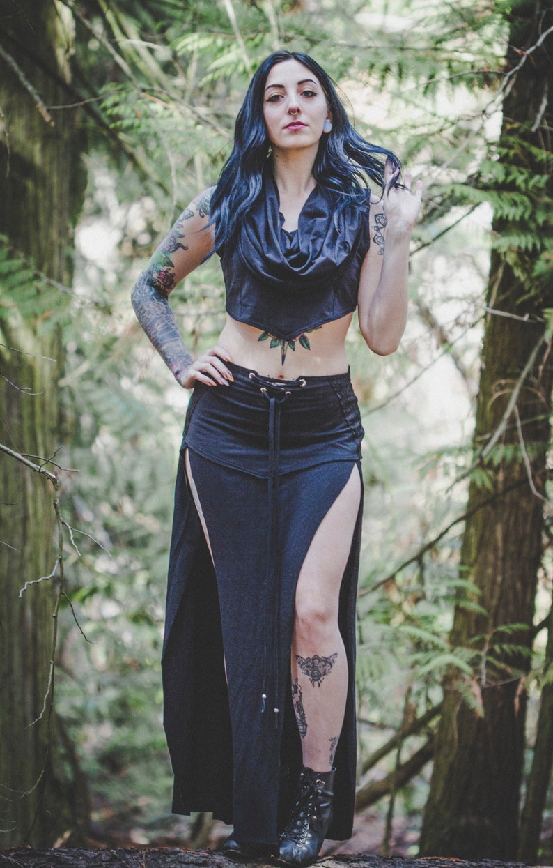 May include: A woman with blue hair and tattoos wears a black crop top with a cowl neck and a long black skirt with side slits. The skirt has a lace-up detail at the waist.